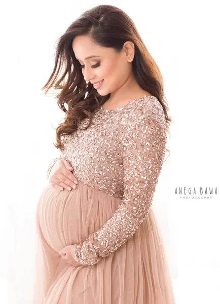 Smiling expecting mother in a dusty rose gown with a sequinned long-sleeve bodice and a tulle skirt, hair blowing, looking down at her