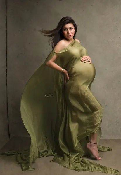Expecting mother in an olive green satin drape slipping off one shoulder with a long train, hair blowing, hand on her bump, textured
