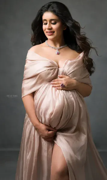 Smiling expecting mother in a blush satin wrap off the shoulders with a jewelled pendant necklace and high slit, hands cradling her bump
