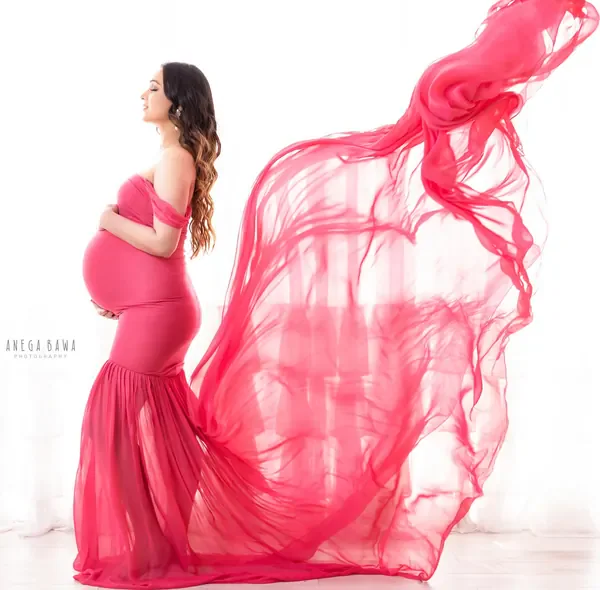 Expecting mother in profile in a hot pink off-shoulder mermaid gown with a long sheer pink train billowing to the side, bright white