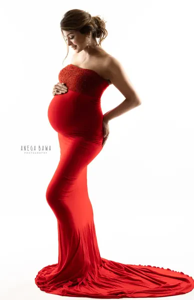 Backlit expecting mother in a fitted red strapless beaded gown with a long train, head tilted down toward her bump, hair in an updo, bright