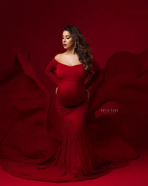 Expecting mother in a red off-shoulder long-sleeve mermaid gown with red fabric flying around her, looking to the side, hand on hip, solid