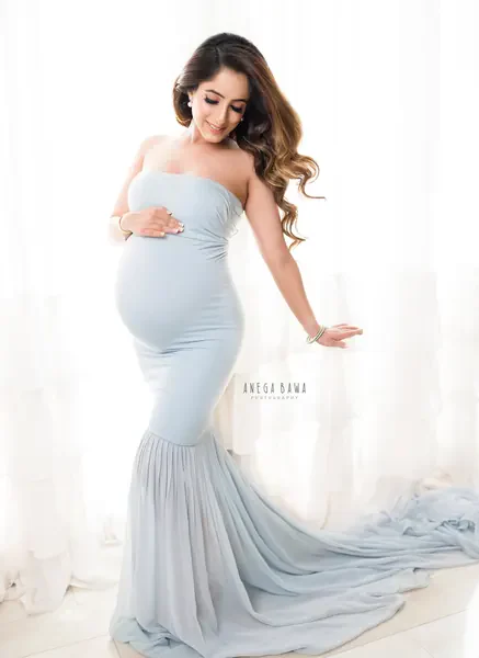 Smiling expecting mother in a pale blue strapless mermaid gown with a long chiffon train, looking down, one hand on her bump and one