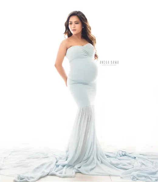 Expecting mother in a pale blue strapless ruched mermaid gown with a long chiffon train spread on the floor, looking to the side, bright