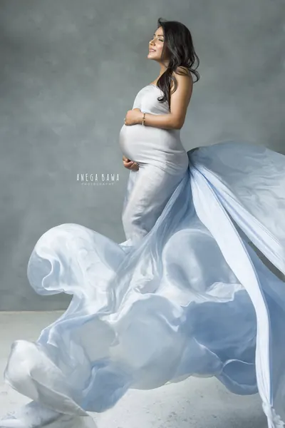 Expecting mother in a fitted white strapless gown with a pale blue silk train billowing out on the floor, looking up, hand on her bump