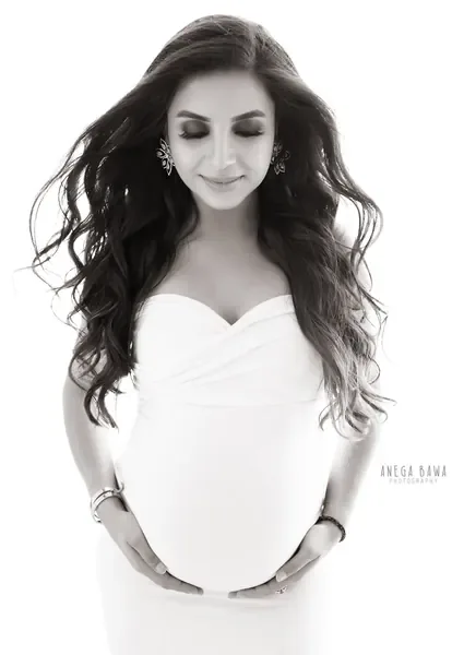High-key black and white portrait of expecting mother in a white sweetheart-neck gown with long wavy hair and drop earrings, eyes closed