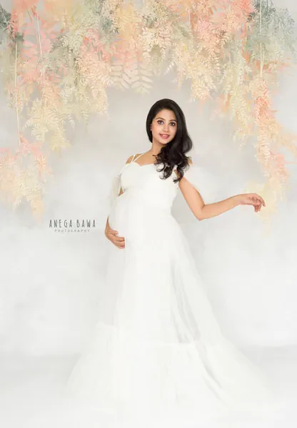 Smiling expecting mother in a white tulle gown with a tiered skirt, one arm out, standing under a hanging peach and cream foliage backdrop.