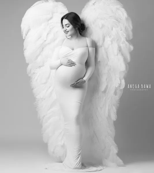 Black and white portrait of expecting mother in a fitted white off-shoulder gown standing in front of large white feather angel wings