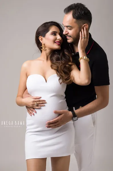 Couple portrait, wife in a white strapless sweetheart mini dress and gold earrings, husband in a black polo and white trousers holding her