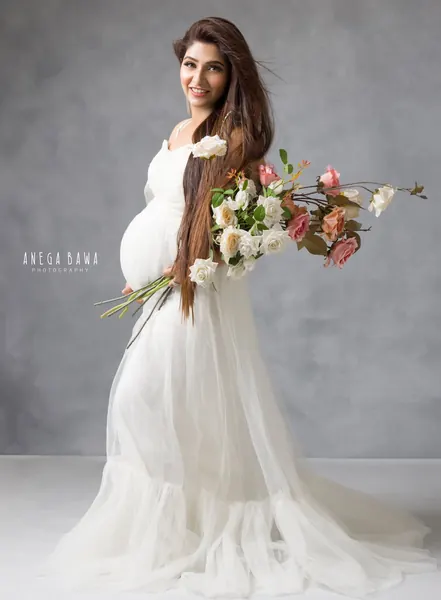 Smiling expecting mother in a white tulle gown with a tiered skirt, long straight hair, holding a trailing bouquet of white and pink roses