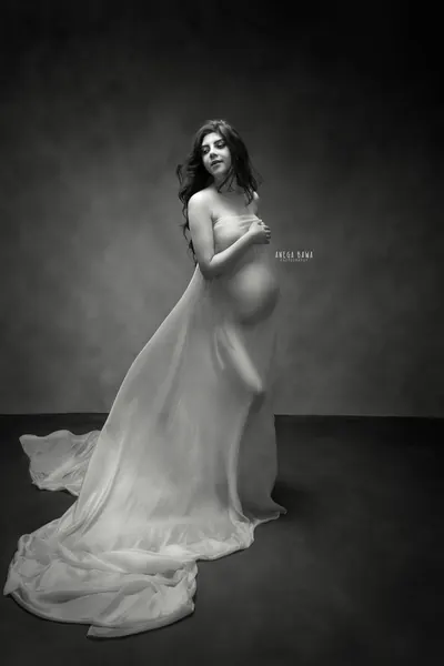 Black and white portrait of expecting mother wrapped in sheer fabric with a long train, bare shoulder, hand across her chest, dark textured