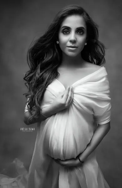 Black and white close portrait of expecting mother with long wavy hair holding a gathered white tulle wrap across her bump, textured grey
