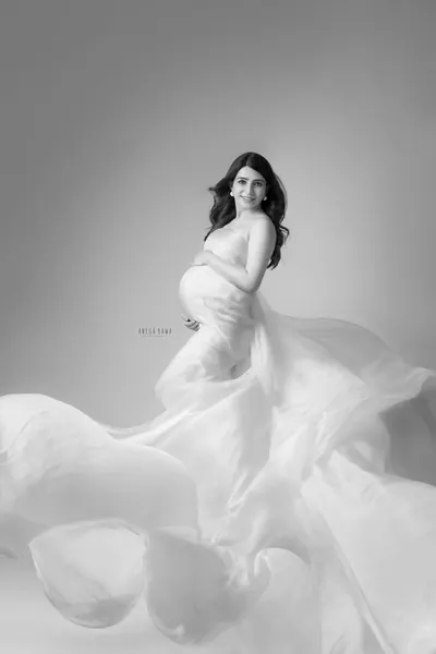 Black and white portrait of smiling expecting mother in a sheer white drape with voluminous flowing fabric at her feet, hand on her bump