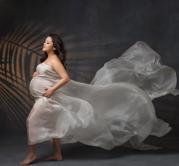 Expecting mother in a pearl-grey satin drape with fabric billowing to the side, barefoot, looking up, palm-leaf shadow on a dark backdrop.