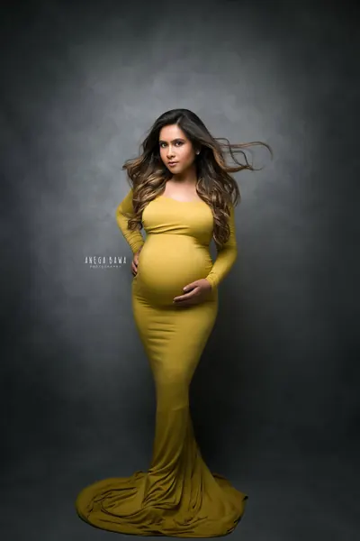 Expecting mother in a mustard long-sleeve fitted gown with a train, hair blowing, hand on hip and one on her bump, dark vignette backdrop.