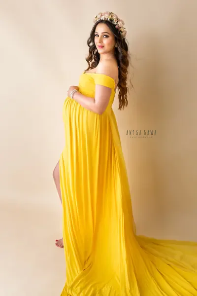 Expecting mother in a bright yellow off-shoulder pleated gown with a high slit and a long train, flower crown, hand on her bump, warm cream