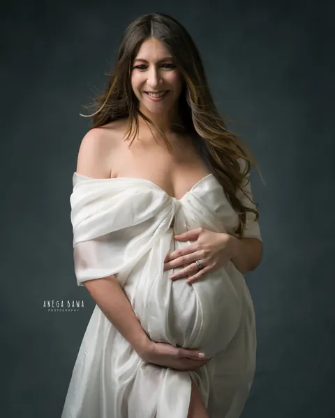 Maternity photography in Delhi: smiling expecting mother in an ivory silk drape tied at the chest, off-shoulder, hands cradling her bump