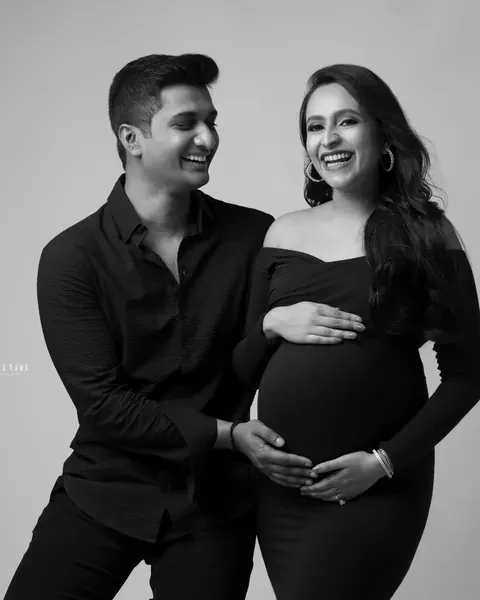 Maternity photography in Delhi: black and white portrait of a laughing couple in black, husband's hands on her bump, grey backdrop, by