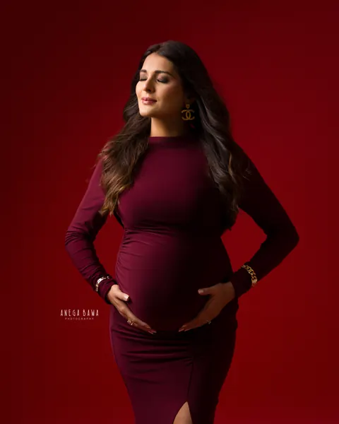 Maternity photography in Delhi: expecting mother in a fitted maroon ruched long-sleeve gown with eyes closed and hands cradling her bump