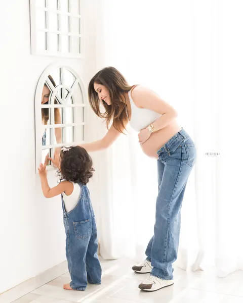 Maternity photography in Delhi: expecting mother in a white top and jeans smiling at her toddler daughter who reaches toward an arched
