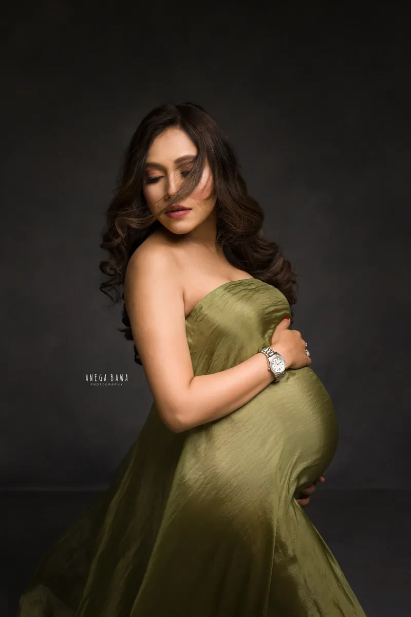 Maternity photography in Delhi: expecting mother in an olive green strapless drape with windswept hair across her face, hands on her bump