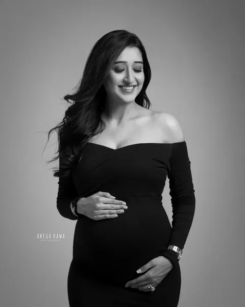 Maternity photography in Delhi: black and white portrait of smiling expecting mother in a fitted black off-shoulder gown, eyes closed