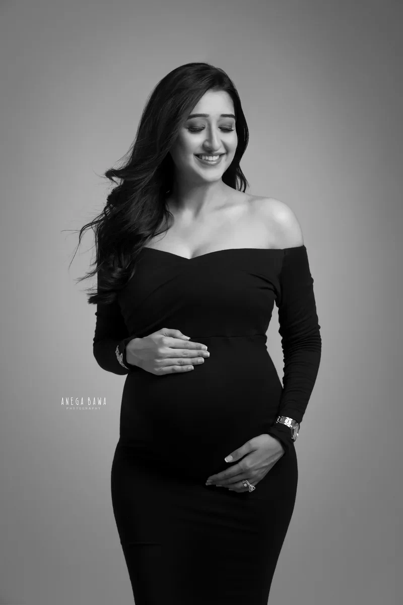 Maternity photography in Delhi: black and white portrait of smiling expecting mother in a fitted black off-shoulder gown, eyes closed