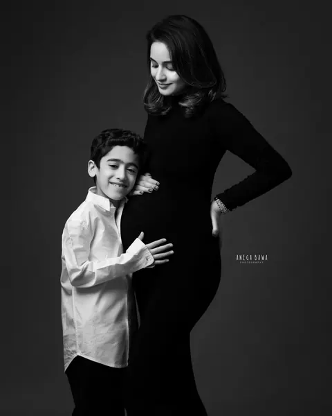 Maternity photography in Delhi: black and white portrait of a smiling boy in a white shirt hugging his mother's bump, she wears a black
