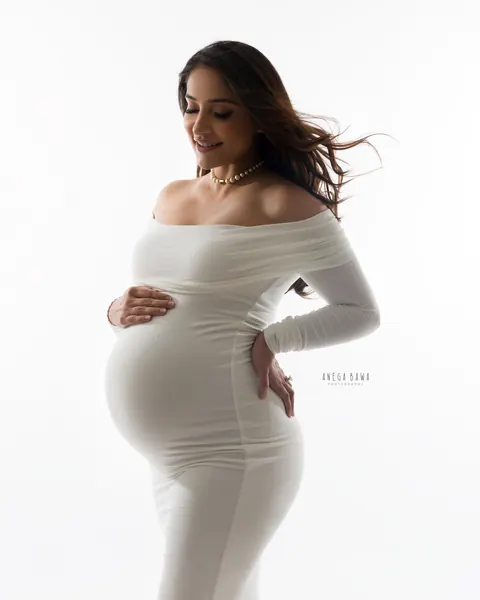 Maternity photography in Delhi: smiling expecting mother in a fitted white off-shoulder gown and pearl necklace with windswept hair, hands