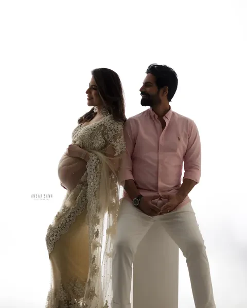 Maternity photography in Gurgaon: backlit couple, wife in an embellished net saree looking away and husband in a pink shirt seated on a