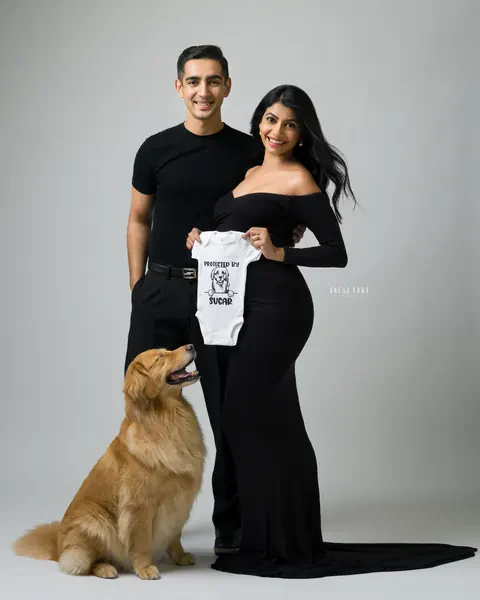 Maternity photography in Gurgaon: couple in black holding a white baby bodysuit, wife in an off-shoulder gown with a train and their golden