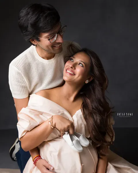 Maternity photography in Gurgaon: smiling couple looking at each other, wife in a champagne gown holding a pair of white baby shoes on her