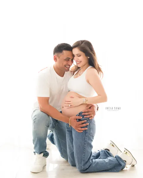 Maternity photography in Gurgaon: couple kneeling together on a white floor in white tops and denim, husband's hands on her bare bump, by