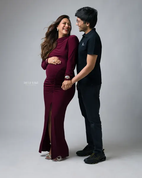 Maternity photography in Gurgaon: laughing couple holding hands, wife in a burgundy ruched gown with a front slit, husband in black, grey