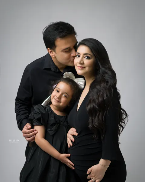 Maternity photography in Gurgaon: husband in a black shirt kissing his wife's cheek while their young daughter in a black dress and bow