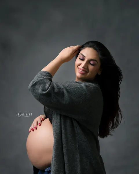 Maternity photography in Gurgaon: smiling expecting mother in an open grey cardigan and jeans with one hand in her hair and the other on