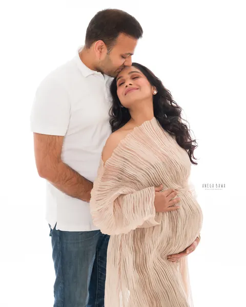 Maternity photography in Gurgaon: husband in a white polo kissing his wife's forehead as she smiles in a blush pleated gown, hands on her