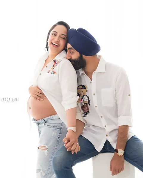 Maternity photography in Gurgaon: laughing wife leaning on her husband who sits on a white block, both in white shirts and denim, bare bump