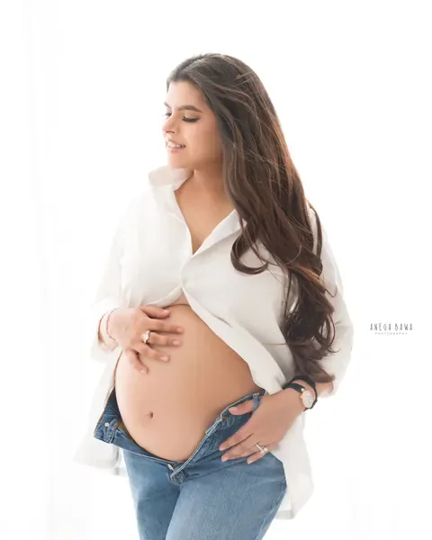 Maternity photography in Gurgaon: expecting mother in an open white shirt and low-rise jeans with a hand on her bare bump, soft window