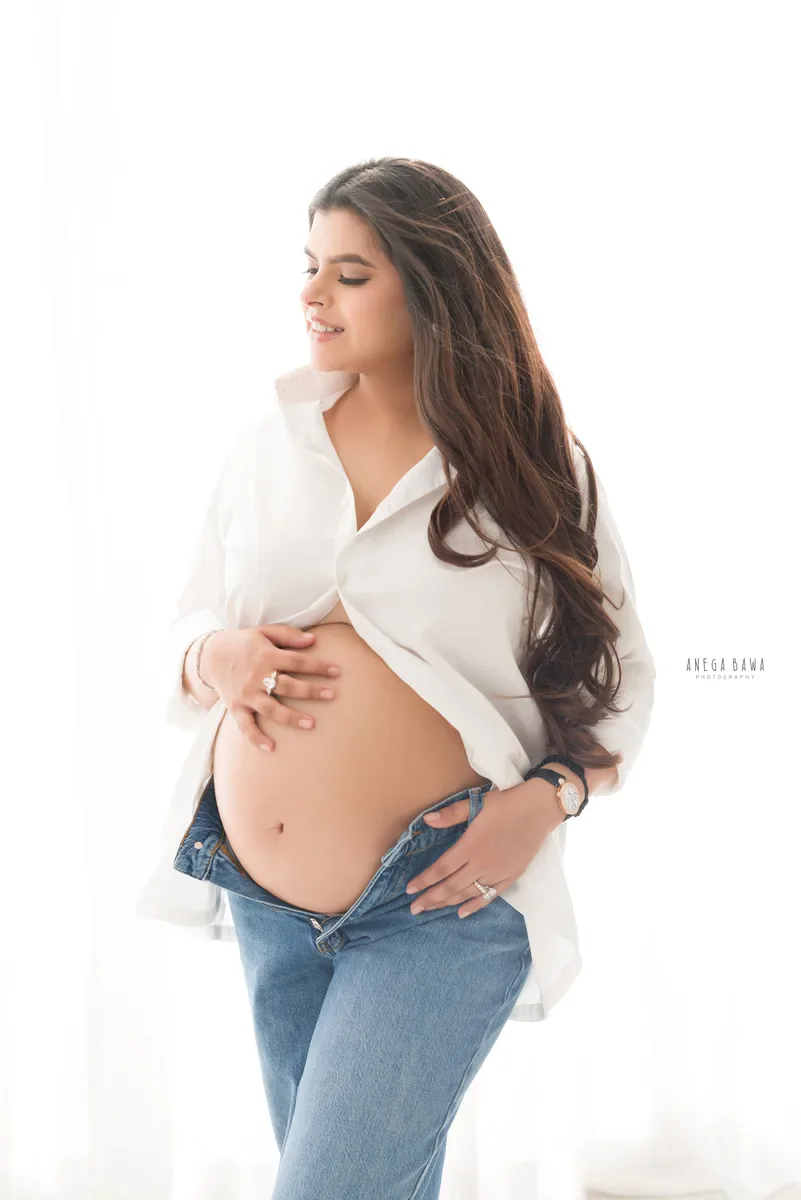 Maternity photography in Gurgaon: expecting mother in an open white shirt and low-rise jeans with a hand on her bare bump, soft window