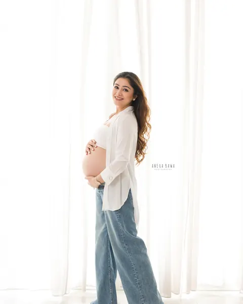 Maternity photography in Gurgaon: smiling expecting mother in an open white shirt and wide-leg denim standing barefoot in front of sheer