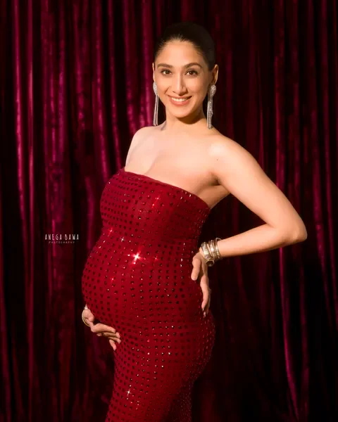 Maternity photography in Gurgaon: smiling expecting mother in a strapless red crystal-studded gown with a hand on her hip, burgundy velvet