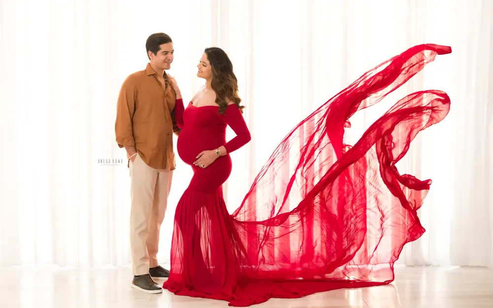 Maternity photography in Gurgaon: expecting mother in a red off-shoulder mermaid gown with a sheer train blown out behind her, husband in a