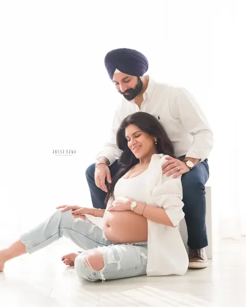 Maternity photography in Gurgaon: expecting mother seated on the floor smiling down at her bump while her husband sits on a white block