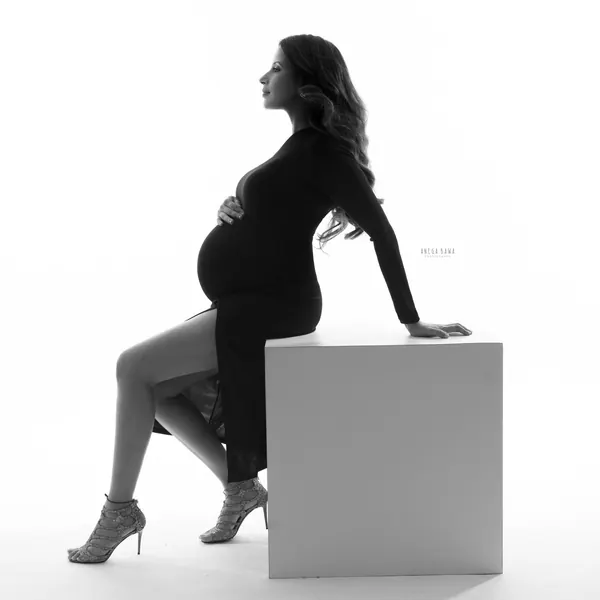 High-key black silhouette of expecting mother seated on a white block in a black long-sleeve dress with a high slit and strappy heels, hand