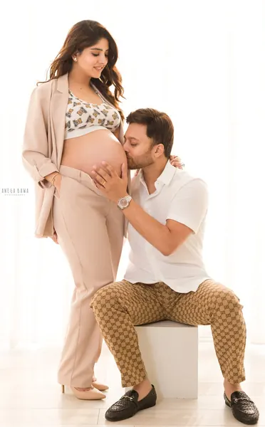 Couple with husband seated on a white block kissing his wife's bare bump, wife in a beige blazer, matching trousers and a patterned crop