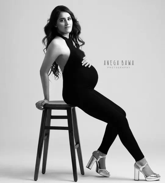 Black and white portrait of expecting mother seated sideways on a dark stool in a black halter jumpsuit and chunky metallic platform heels
