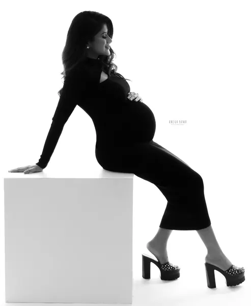 High-key black silhouette of expecting mother seated on a white block in a black fitted long-sleeve dress and studded platform heels, hand