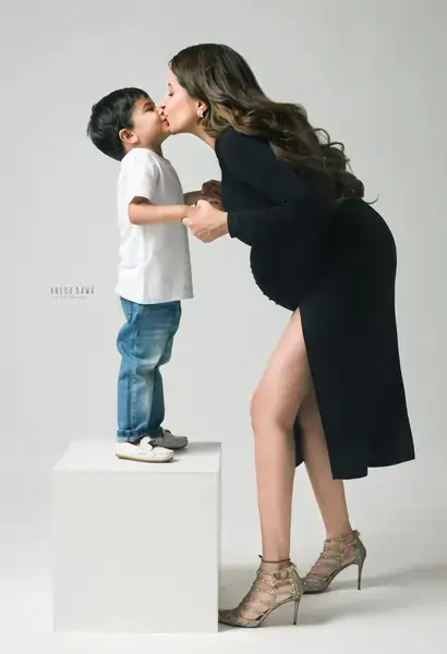 Young son in white tee and jeans standing on a white block to kiss his expecting mother, who wears a black wrap dress with a high slit and