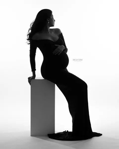Maternity photoshoot in Delhi: black and white silhouette of expecting mother in an off-shoulder gown seated on a white block, looking over